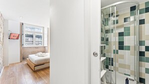 Et hotelværelse med seng, vindue, bruser med tern og et tv ophængt på væggen.