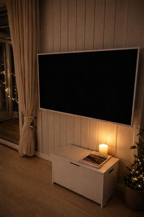 A modern room with a large flat-screen TV mounted on a wall, a bedside table with a book and a candle, a window with curtains, and a potted plant.