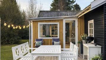 Ett trädäck med ett vitt bord och bänkar, en pergola med ljusslingor och en grillplats.