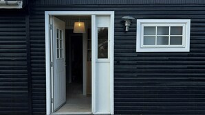 A black building with a white door and window, a wooden deck, and a light fixture above the door. - Little oasis with large terrace and a cozy garden (Grevinge)