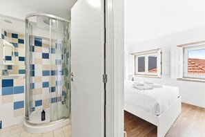 A bathroom with a glass shower enclosure and a tiled floor, a bedroom with a bed and a window, and a white door leading to another room. - GuestReady - InPorto Building - Room 6 (Porto)