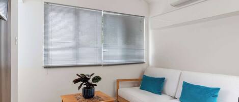 A room with a white sofa, wooden floor, a small table with a plant, and a window with blinds.