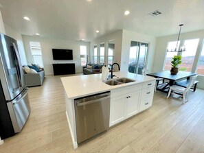 A modern kitchen with a dishwasher, a dining area with a table and benches, and a living area with a sofa and TV. - Paseos Pool Oasis | Outdoor Pool & Hot Tub (Washington)