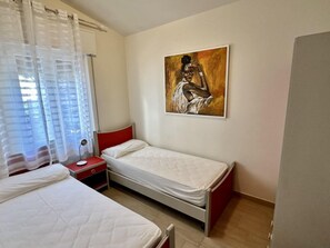 A bedroom with two beds, a nightstand, a painting on the wall, and a window with sheer curtains. - Seafront Flat With Stunning Views (San Michele al Tagliamento)