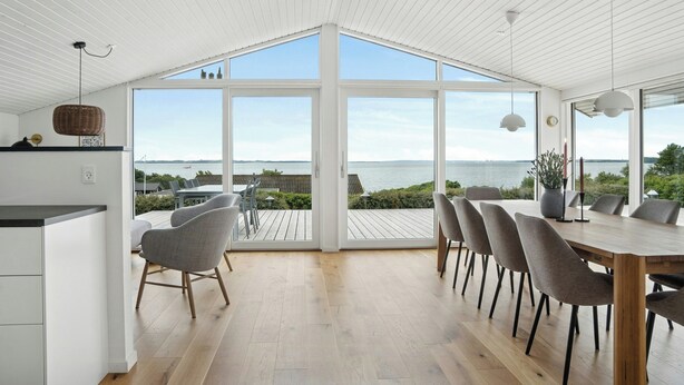 Un espace de restauration moderne doté d’une grande table et de chaises, offrant une vue sur la mer, et présentant un intérieur blanc avec un revêtement de sol en bois.