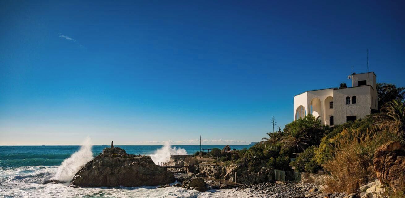 Un bâtiment situé sur la côte, doté de fenêtres en arc de cercle, qui se trouve sur un littoral rocheux où les vagues s’écrasent contre les rochers.