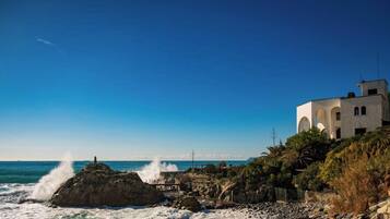 Un bâtiment situé sur la côte, doté de fenêtres en arc de cercle, qui se trouve sur un littoral rocheux où les vagues s’écrasent contre les rochers.