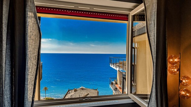 Habitación con un amplio ventanal con vistas a la costa, un balcón con barandilla y un aplique de pared.