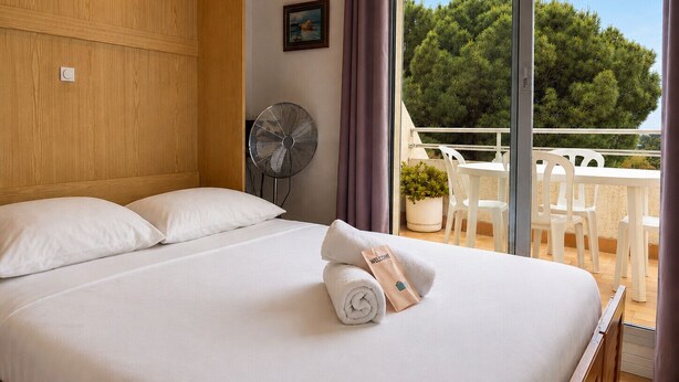 Ein Hotelzimmer mit Bett, einem Ventilator, einer Willkommensauslage und einem Balkon mit Blick auf Bäume.