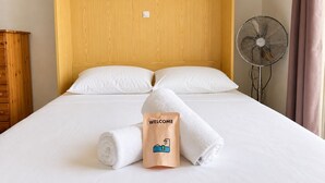 Ein Hotelzimmer mit Holz-Kopfteil, zwei weiße Kissen, einem Bett mit weißer Bettwäsche, einem zusammengerollten weißen Handtuch, einem Ventilator und einer Willkommensausgabe.