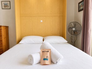 Ein Hotelzimmer mit Holz-Kopfteil, zwei weiße Kissen, einem Bett mit weißer Bettwäsche, einem zusammengerollten weißen Handtuch, einem Ventilator und einer Willkommensausgabe.