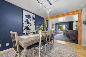Ein Essbereich mit Holztisch und -stühlen, einem großen abstrakten Gemälde an der Wand und Blick in ein Wohnzimmer mit einer Couch und einer Tür, die nach draußen führt.
