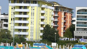 Edifici de diverses plantes amb balcons, zona de piscina amb ombrel·les i un cel serè.