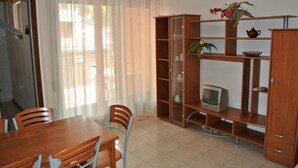 Una habitació amb taula de menjador de fusta, cadires, televisió sobre una estant i una finestra amb cortines translúcides.