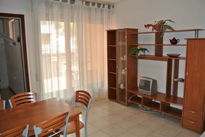 Ein Zimmer mit einem Holzesstisch, Stühlen, einem Fernseher auf einem Regal und einem Fenster mit durchsichtigen Vorhängen.
