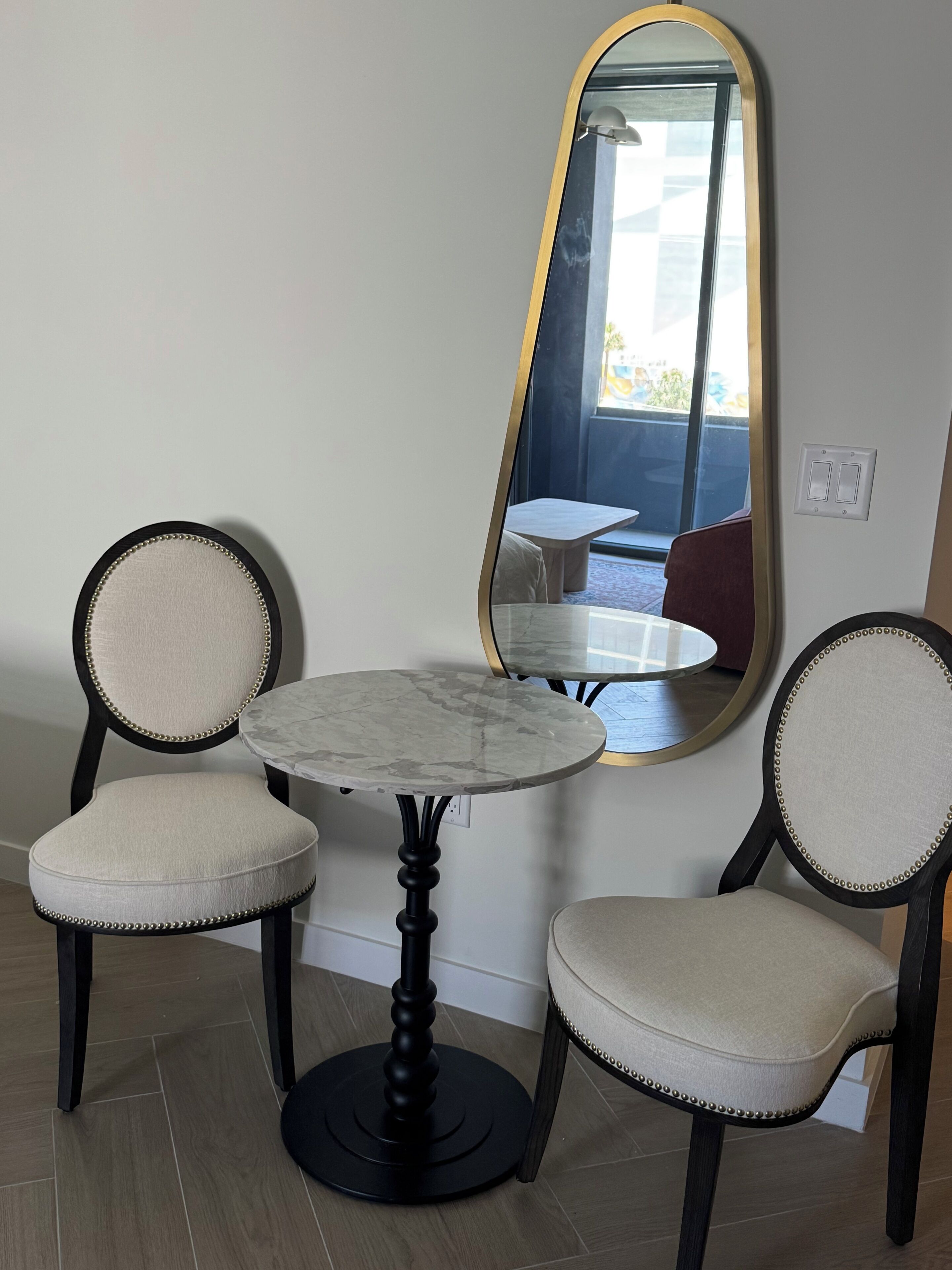 A room with a round table, two upholstered chairs, a large mirror, and a window.