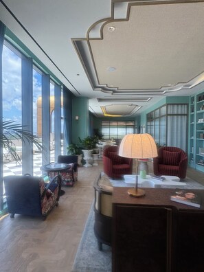 Eine geräumige Lobby mit Schachbrettmuster-Boden, großen Fenstern, einer zentralen Sitzecke mit Tisch und Stühlen sowie einer dekorativen Decke.