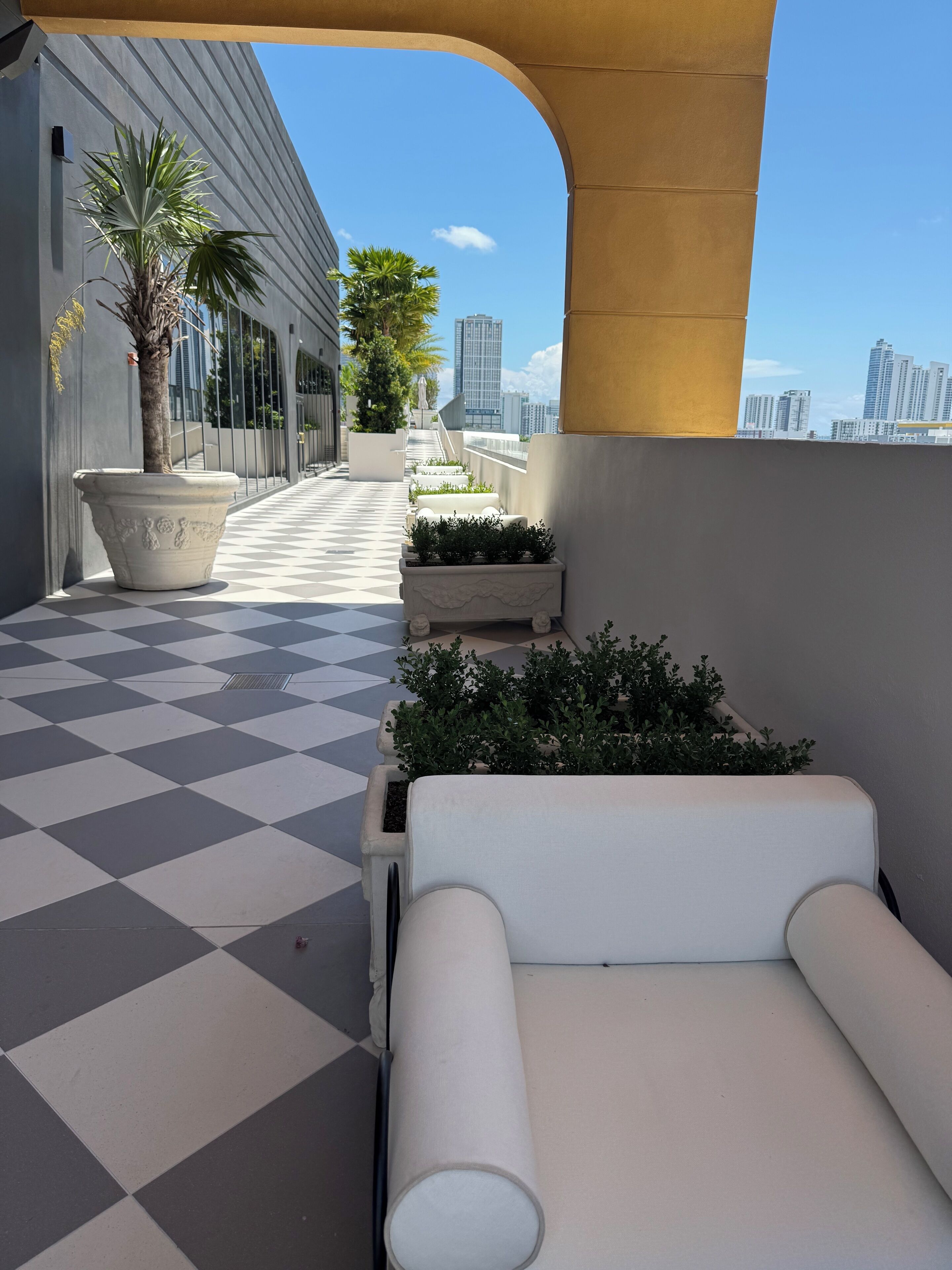 Terraço no telhado com chão xadrez, plantas em vasos, uma cadeira de descanso branca e vista para edifícios da cidade.
