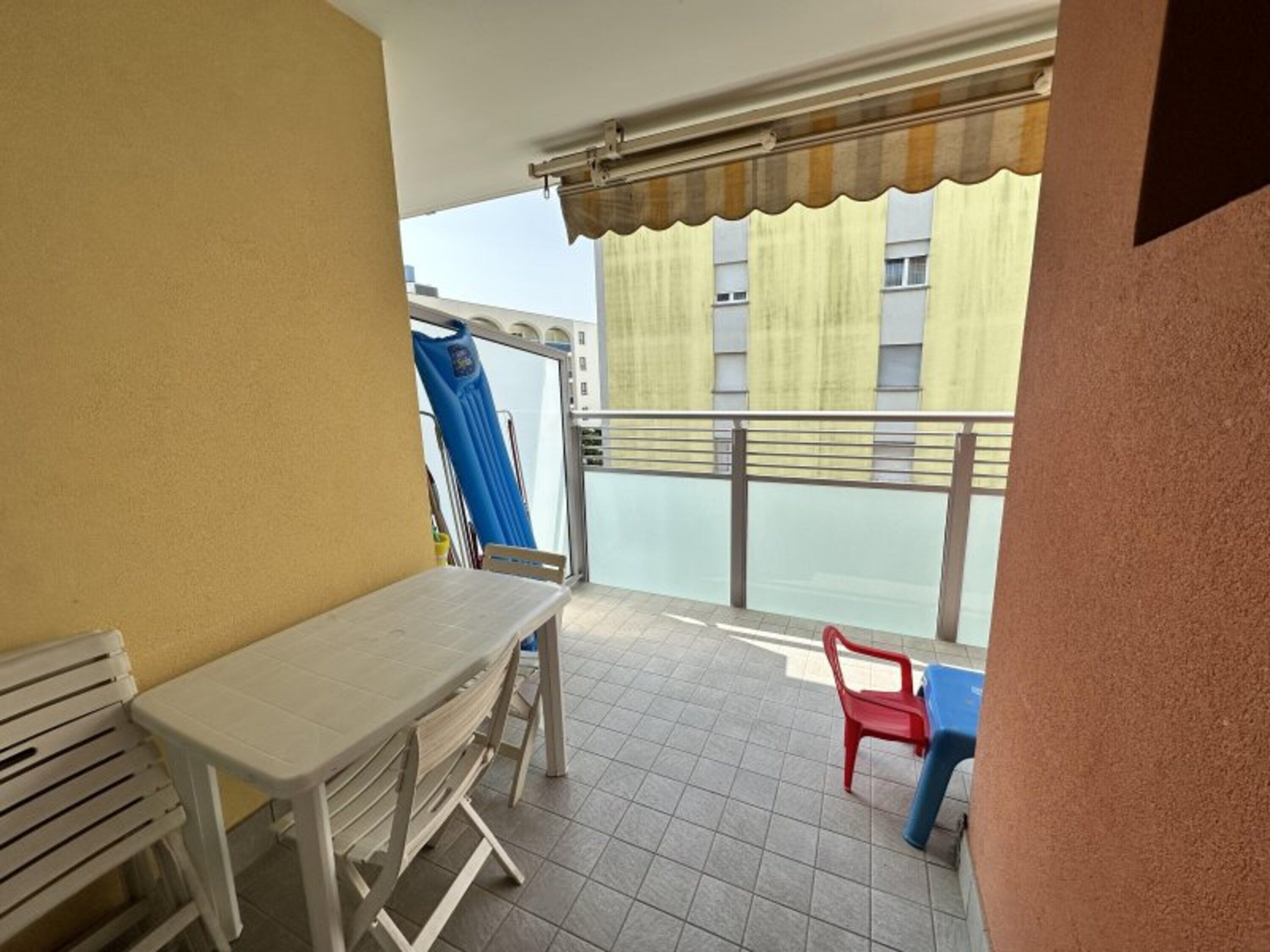 Ein Balkon mit Tisch und Stühlen, ein blauer Stuhl, ein roter Stuhl und ein Fenster mit Vorhängen.