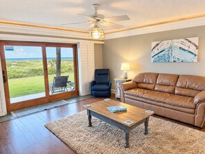 A living room with a sliding glass door leading to a grassy outdoor area, a ceiling fan, a brown leather sofa, a wooden coffee table, and a lamp on a side table. - Sea Haven 218 - Ground Floor, Pool, Beach Access, Putt Putt, Shuffleboard (St. Augustine)