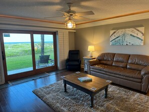 Ein Wohnzimmer mit brauner Ledersofa, einem Holztisch, einem schwarzen Sessel, einem Beistelltisch mit Lampe und Blick auf das Meer durch Glastüren.