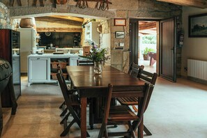 Een rustieke eethoek met een houten tafel en stoelen, een stenen muur en een keuken op de achtergrond.