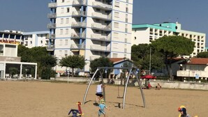 A multi-story building with balconies, a sandy beach area with playground equipment, and a clear sky. - Seafront Flat With Splendid Views From the Terrace (San Michele al Tagliamento)