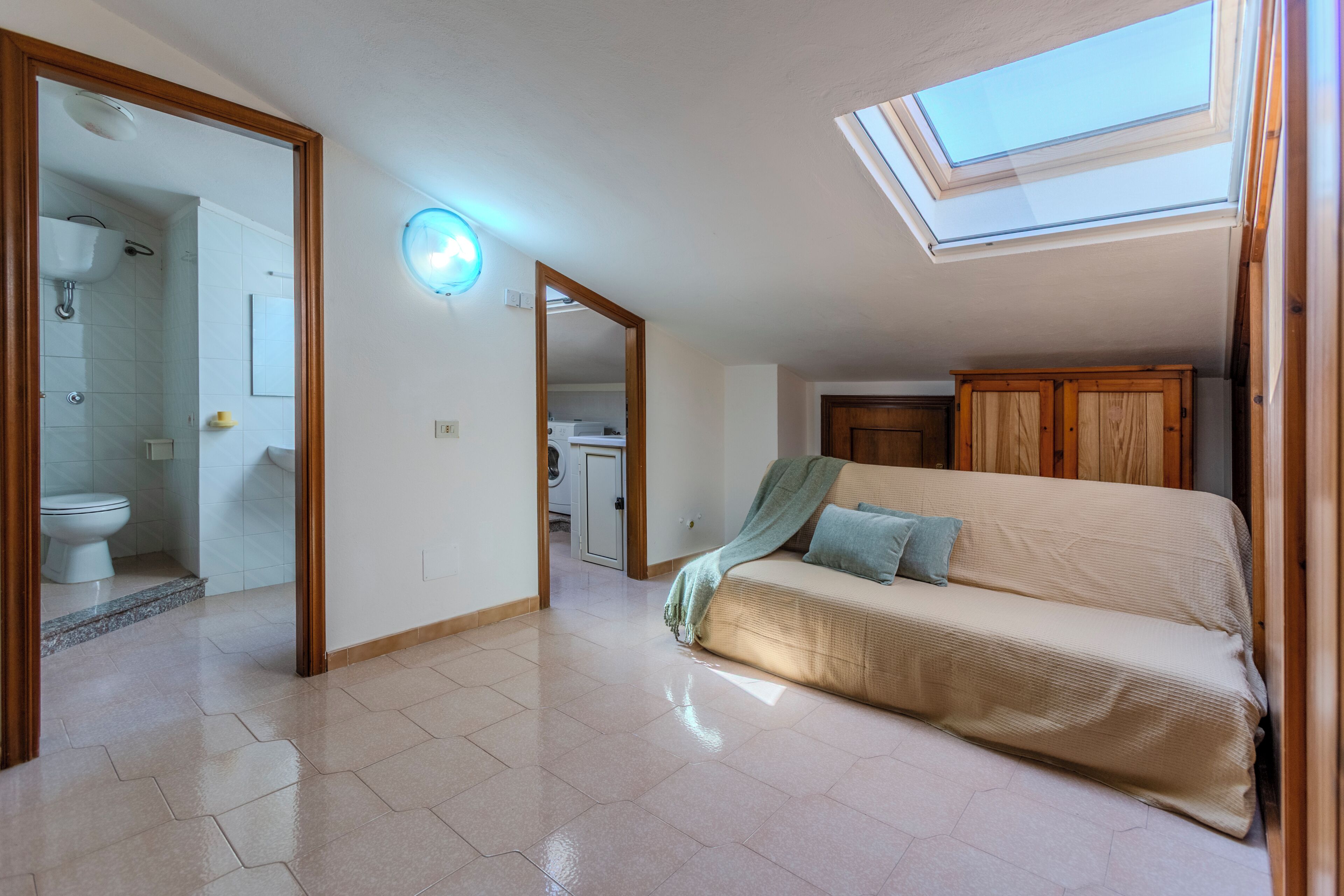 Una stanza con un lucernario, un divano, un armadio in legno e un bagno visibile attraverso una porta aperta.