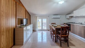 Une cuisine moderne avec des meubles en bois, un coin repas avec des chaises, un téléviseur posé sur un meuble et une vue à travers une porte vitrée.