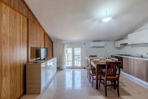 A modern kitchen with wooden cabinets, a dining area with chairs, a TV on a stand, and a view through a glass door.