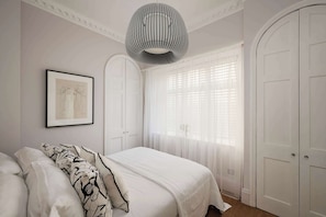 Ein Schlafzimmer mit einem Bett, einem gerahmten Bild, einer Pendelleuchte und einem Fenster mit durchscheinenden Vorhängen.