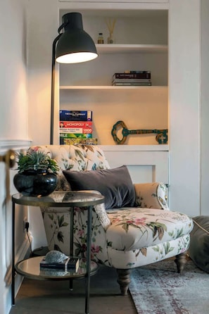 A cozy living room with a floral sofa, a small side table with a plant, a bookshelf with books, and a decorative wall hanging. - The Paragon Abode Bath Central Period Apartment (Bath)