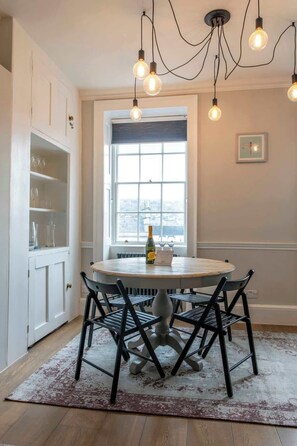 A dining area with a round table, black chairs, a bottle of wine, a menu, and a window with a view. - The Paragon Abode Bath Central Period Apartment (Bath)