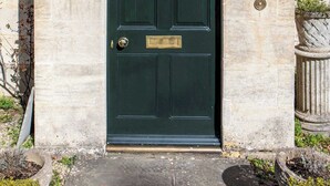 Une porte verte ornée d’un heurtoir en laiton, un mur de pierre et une fenêtre avec des rideaux blancs.