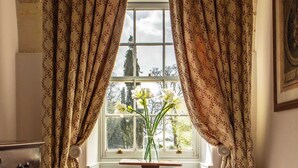 Une pièce avec une fenêtre, une commode, un vase de fleurs et un tapis à motifs.