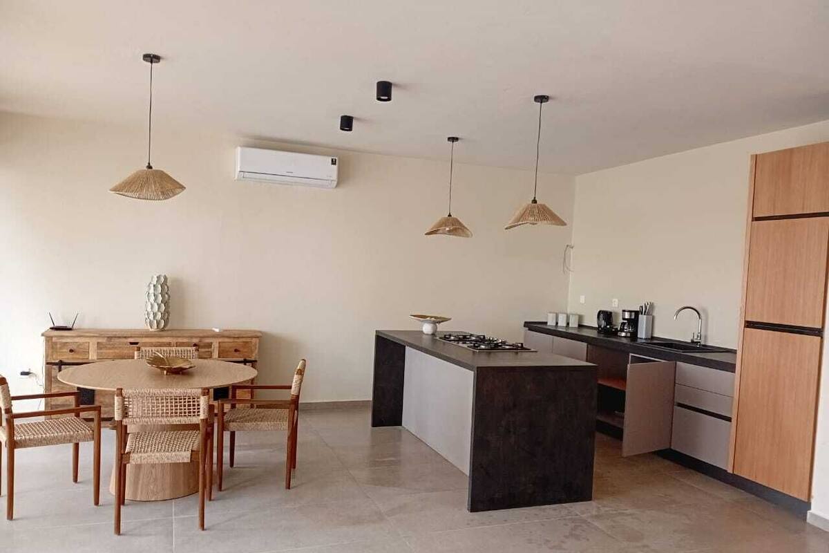 A modern kitchen with a dining area, featuring a round table, chairs, pendant lights, and a built-in island with a stove.