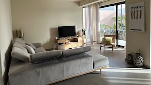 A modern living room with a grey sofa, a flat-screen TV, a wooden console table, and a large window with a view of greenery.