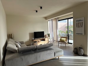 A modern living room with a grey sofa, a flat-screen TV, a wooden console table, and a large window with a view of greenery.