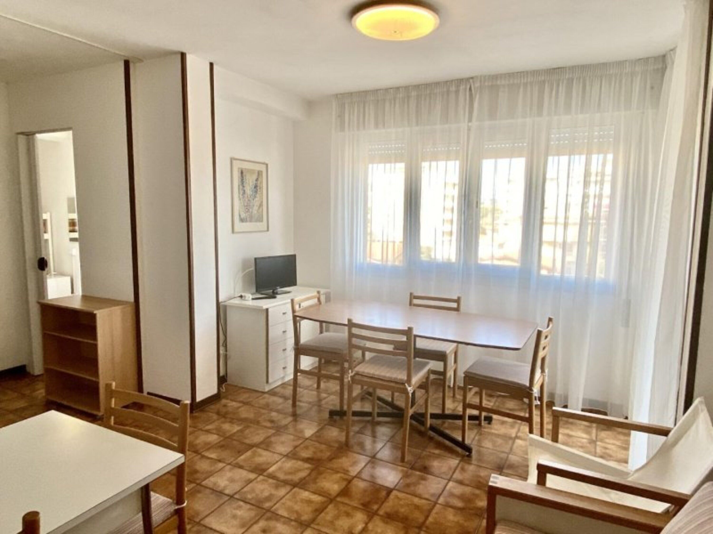 A room with a dining table, chairs, a TV, and a window with sheer curtains.