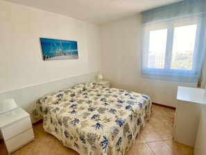 Ein Schlafzimmer mit einem Bett, einem Nachttisch, einem Schreibtisch, einem Fenster und einem Bild an der Wand.