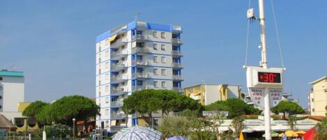 Una playa con un edificio alto, un indicador digital de temperatura y un cartel que muestra 'MAIA' y 'AURIGA', indicando lugares cercanos.