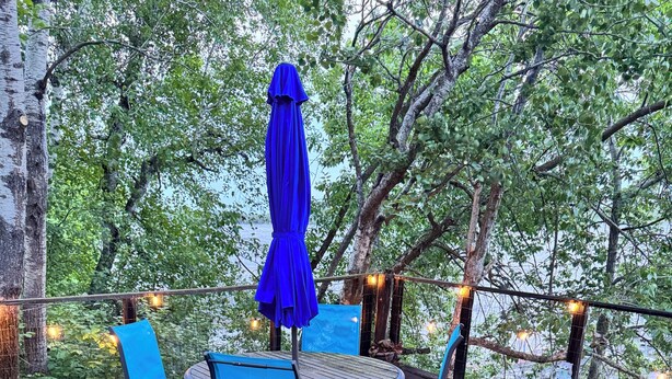 A wooden deck with a round table and blue chairs, surrounded by trees and string lights. - Escape to Lake Metigoshe This Summer (Bottineau)