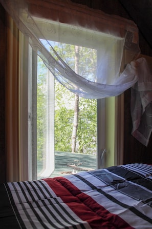 Ein Bett mit gestreifter Bettdecke, ein Fenster mit leichtem Vorhang und Blick auf Bäume.