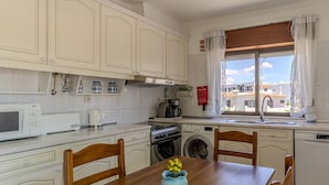 Una cocina con armarios blancos, microondas, estufa, cafetera y ventana con cortinas.