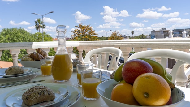 Mesa de desayuno con croissants, jugo y fruta en un balcón con cielo despejado.