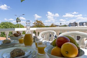 Mesa de café da manhã com croissants, suco e frutas em uma varanda com céu claro.