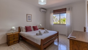 Un dormitorio con una cama de madera, una mesita de noche con lámpara, una ventana con cortinas y vistas a palmeras.