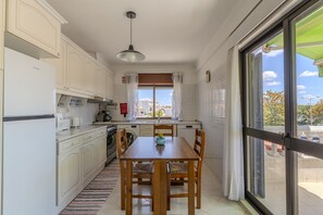 Uma cozinha com armários brancos, uma mesa de jantar com cadeiras, uma janela com cortinas e vista para o exterior.
