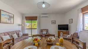 Un comedor con una mesa redonda de madera, sillas, un sofá, un televisor y una ventana con cortinas.