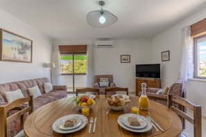 Uma sala de jantar com uma mesa de madeira redonda, cadeiras, um sofá, uma televisão e uma janela com cortinas.